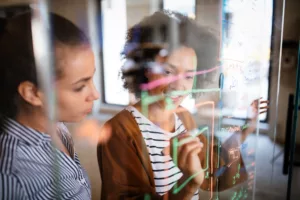 Two women collaborating and writing on a glass wall with markers, with a focus on the woman in the foreground smiling.