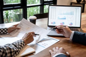 Two professionals analyzing financial data with documents and a laptop displaying graphs.